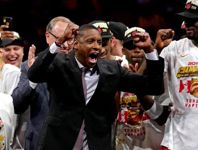 Masai Ujiri raises his fists in celebration of the NBA Championship win, surrounded by members of the Toronto Raptors team.